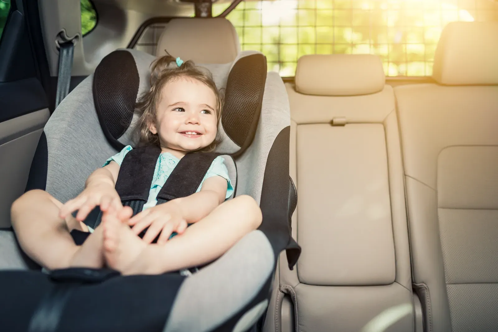 A smiling toddler buckled into a child seat in a beige leather back seat