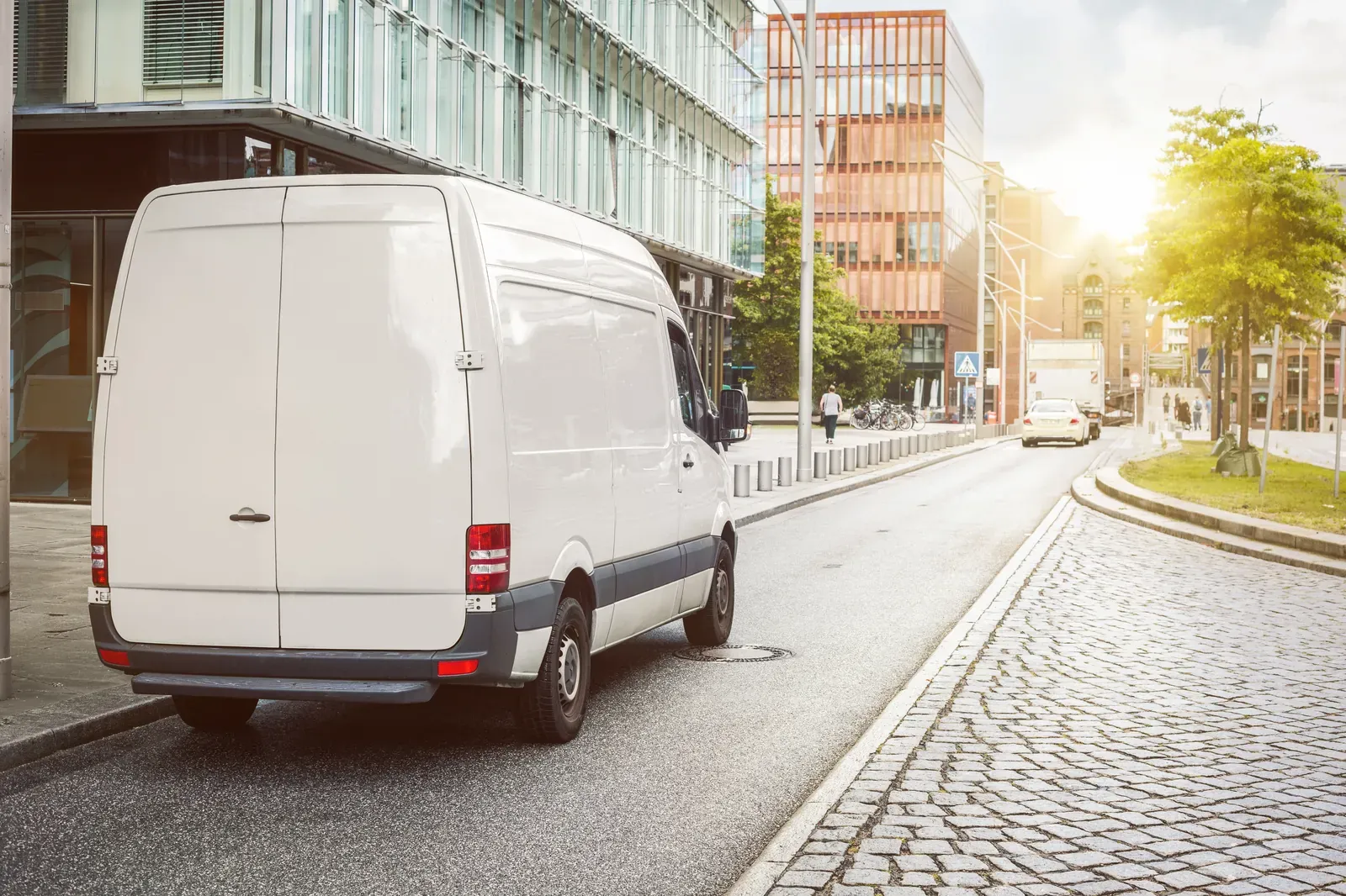 A white cargo van parked on a cobblestone street among modern glass buildings