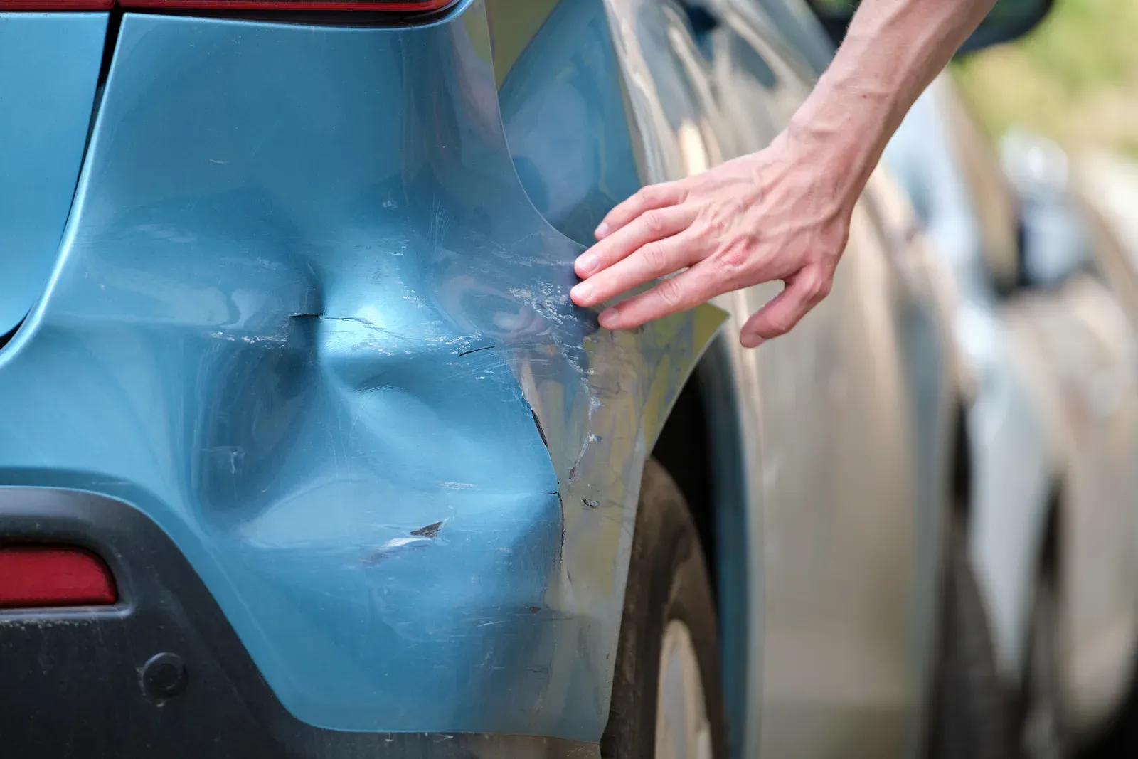 A hand touching the dented, scraped corner of a blue rear bumper