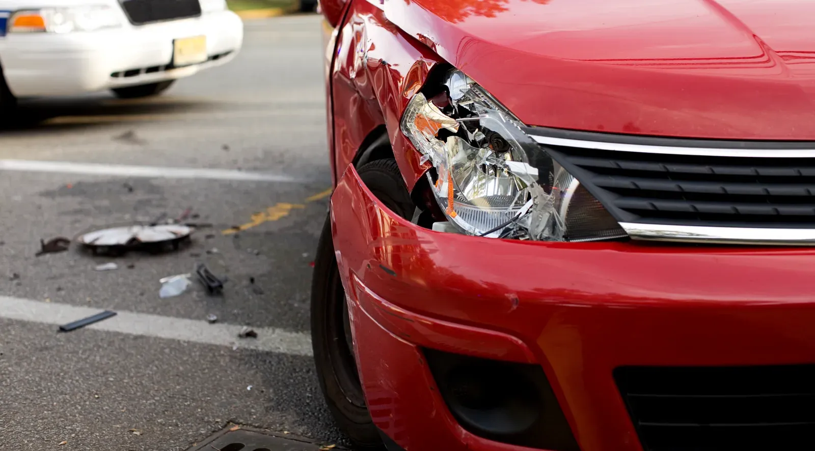 A red car with a shattered headlight and crumpled fender, debris on the road