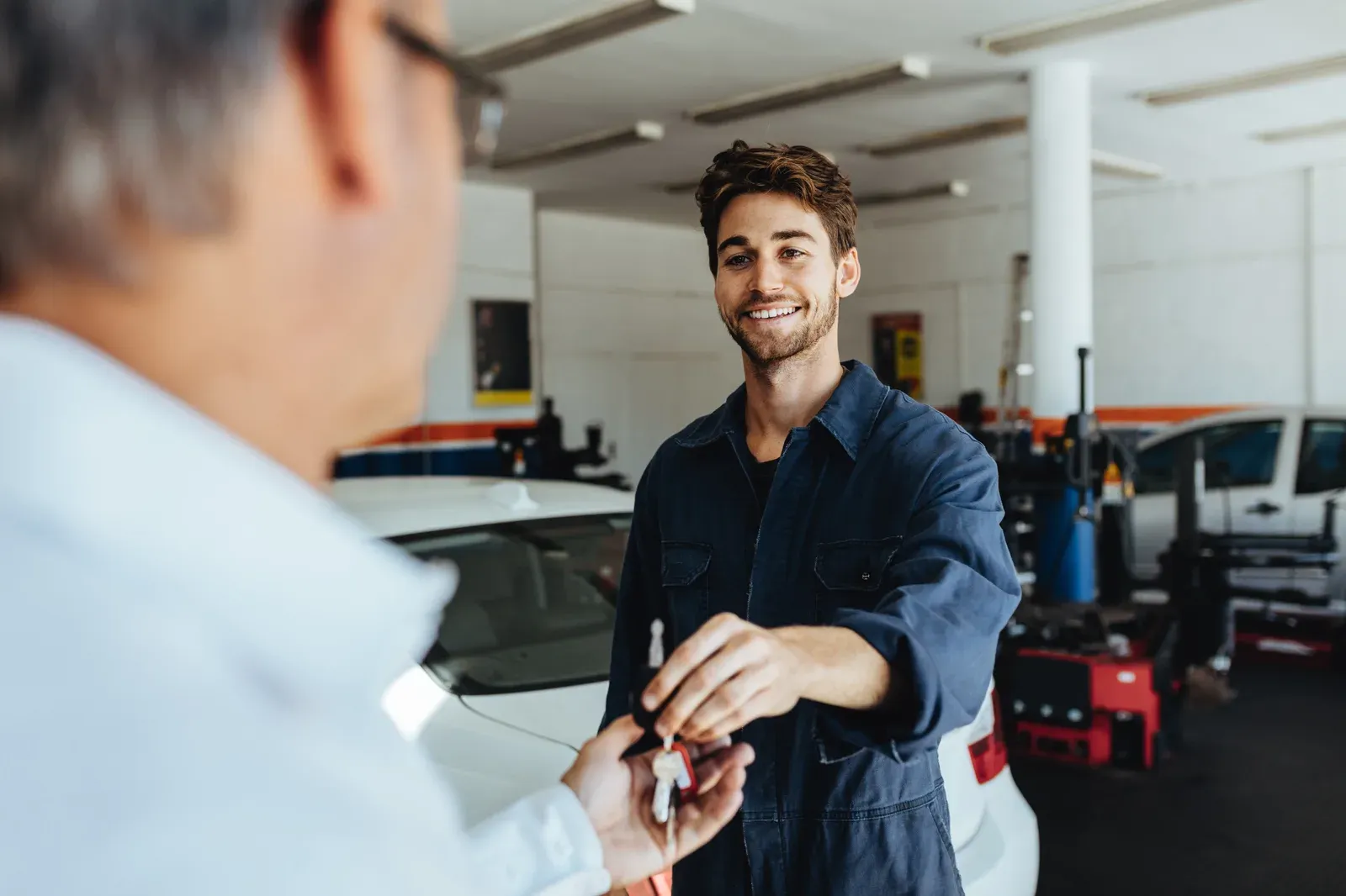 A smiling technician in navy coveralls receiving car keys in a bright workshop