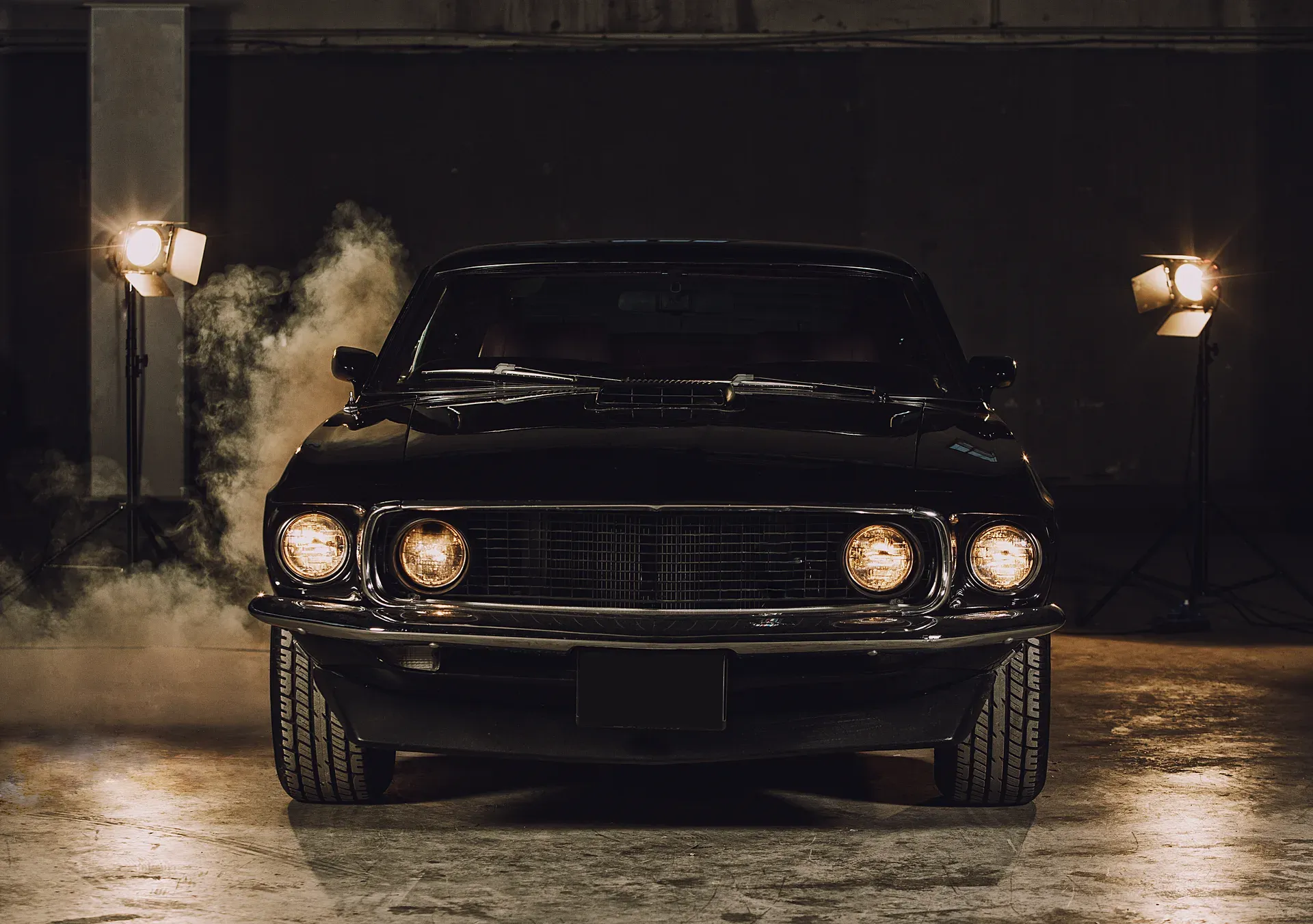 A black classic muscle car with lit headlights in a smoky studio with stage lights