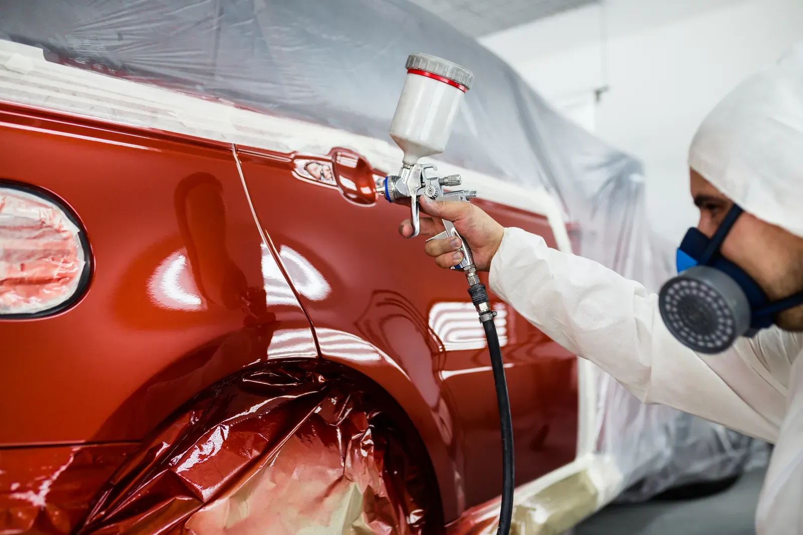 A painter spraying deep red paint onto a masked rear quarter panel