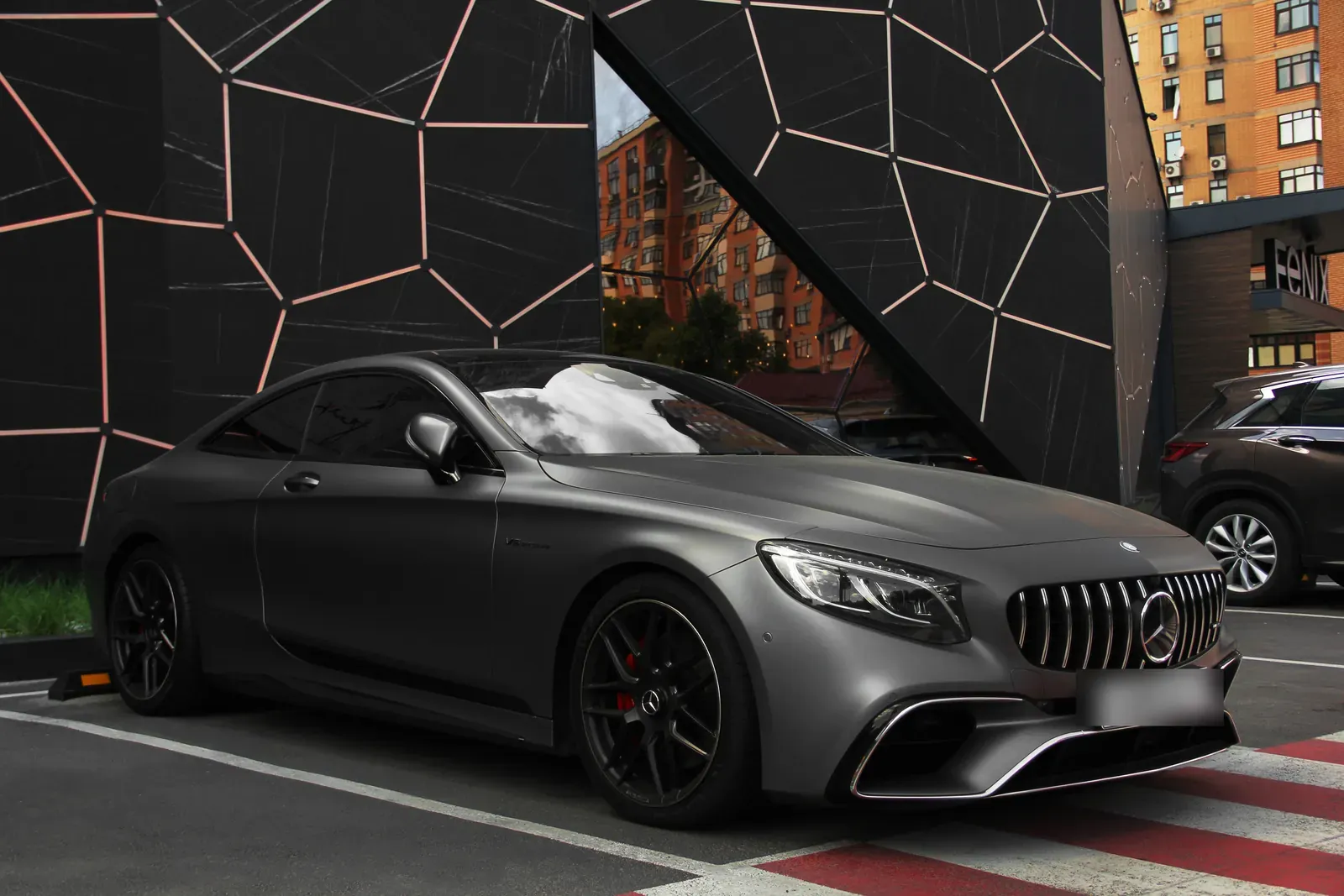 A matte grey coupe parked before a black wall with a geometric line pattern