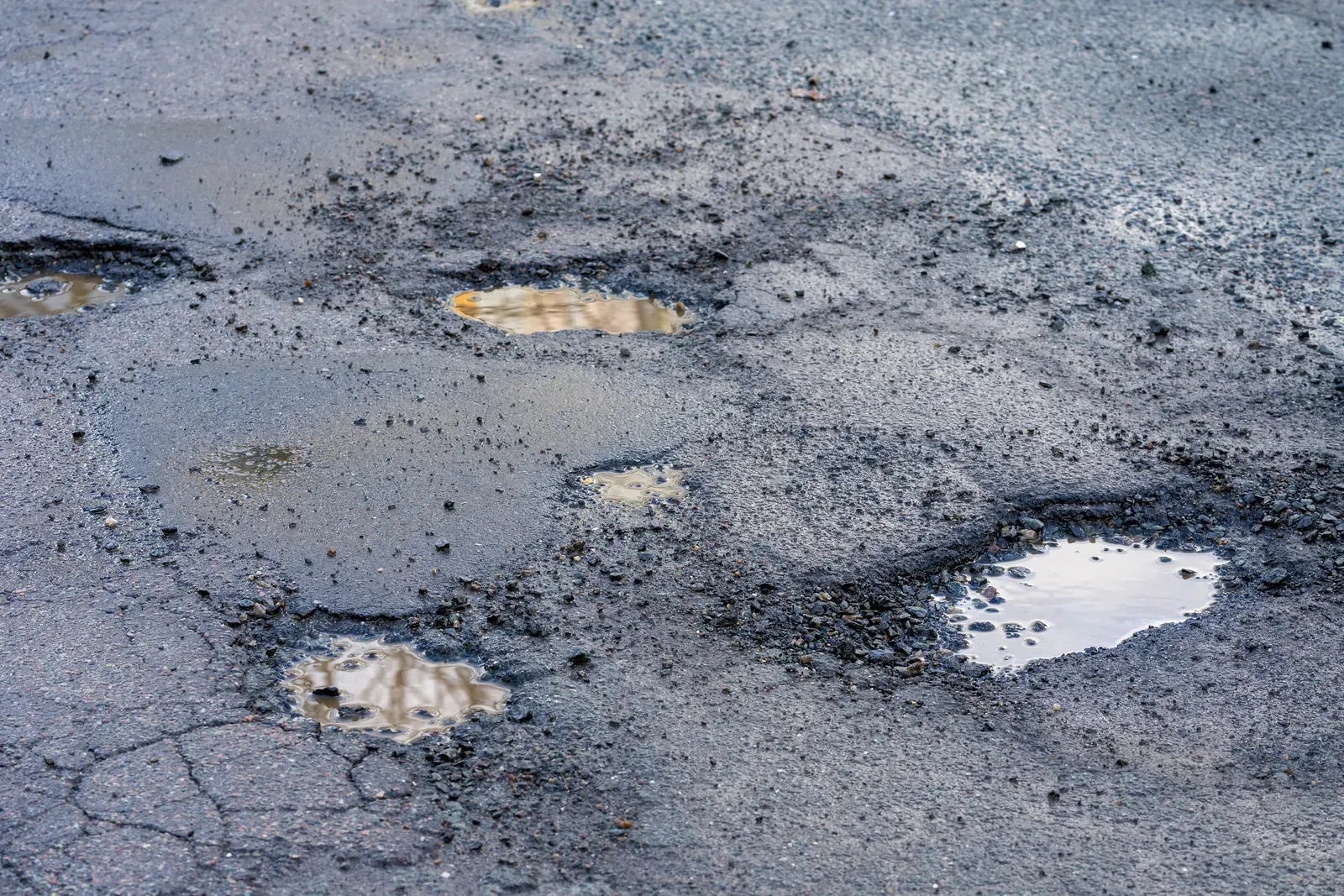 An asphalt road surface broken by several water-filled potholes and loose gravel