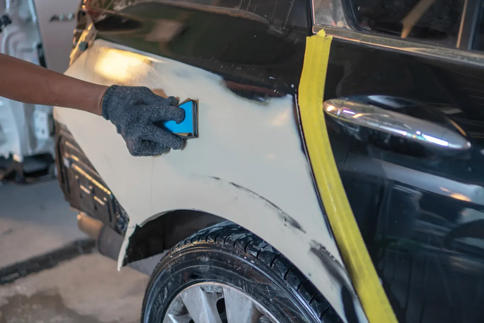 A gloved hand sanding primer on a rear quarter panel masked with yellow tape
