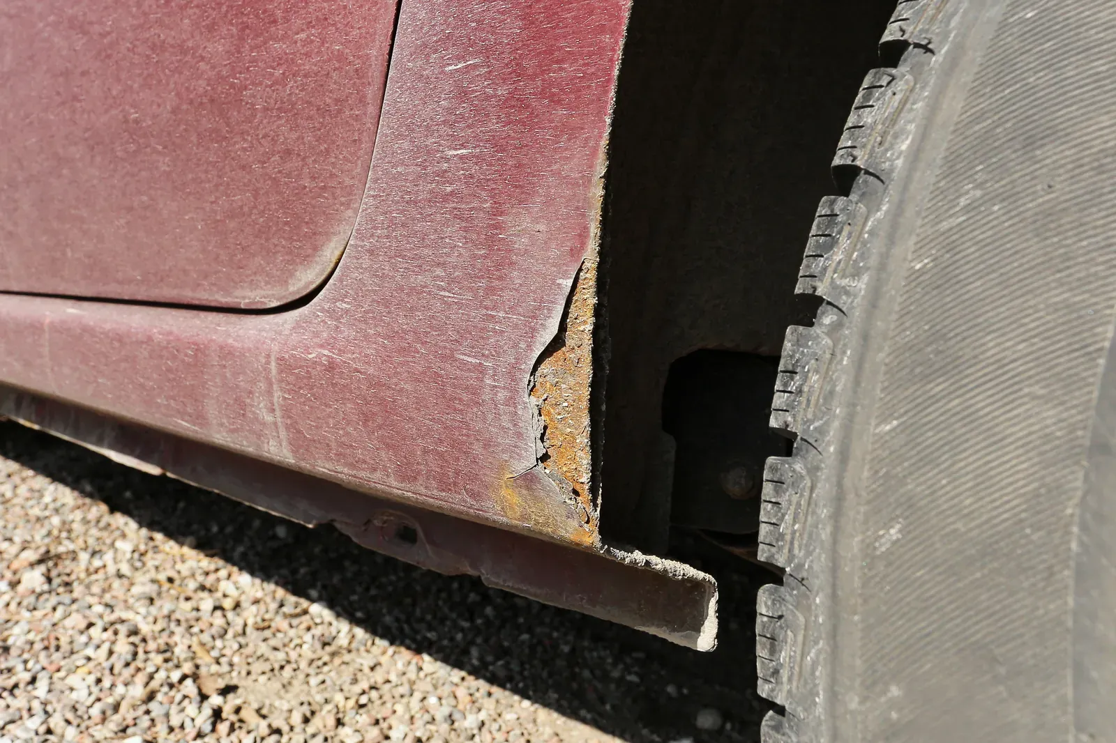 A rusted rocker panel edge crumbling below a dusty maroon door