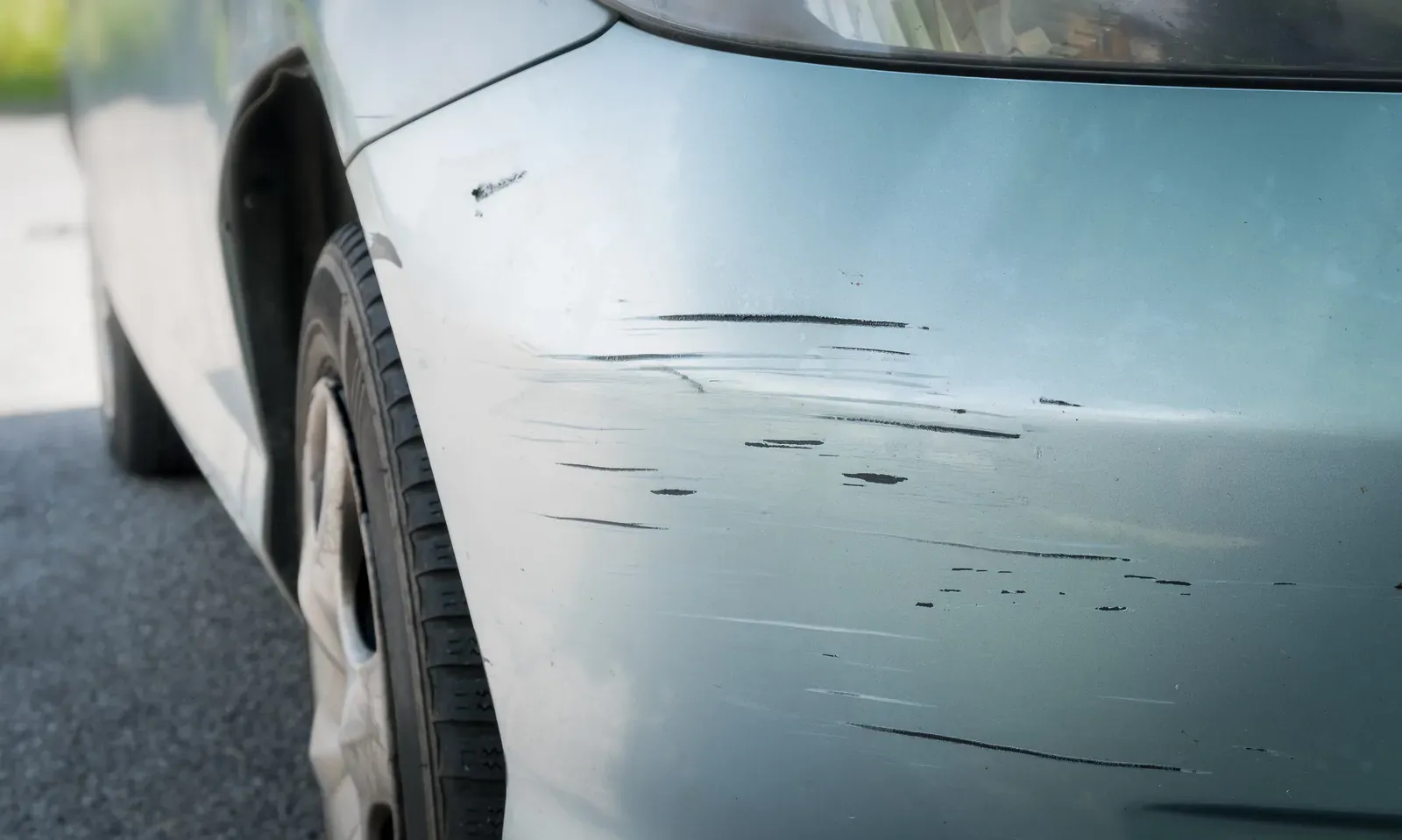 Multiple scratches marring a pale blue front bumper near the headlight