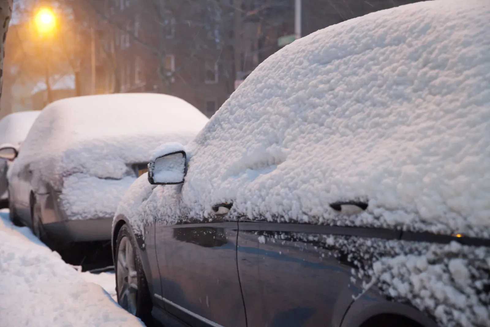 Parked cars buried under thick snow on a street at dusk
