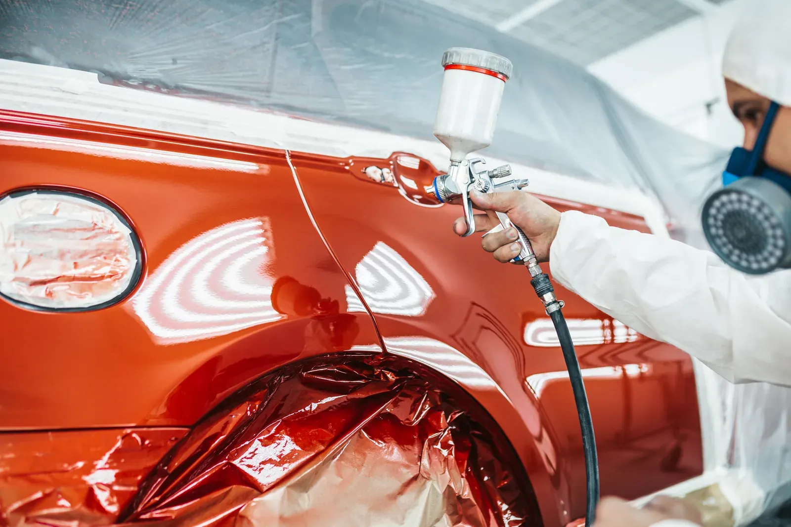 A hand guiding a spray gun along a glossy red quarter panel in a booth
