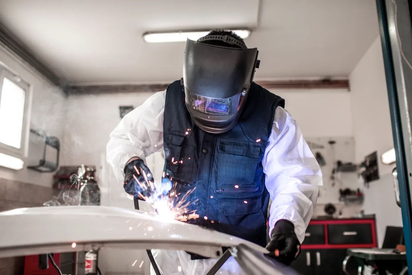 A welder in a helmet joining a white body panel amid sparks and smoke