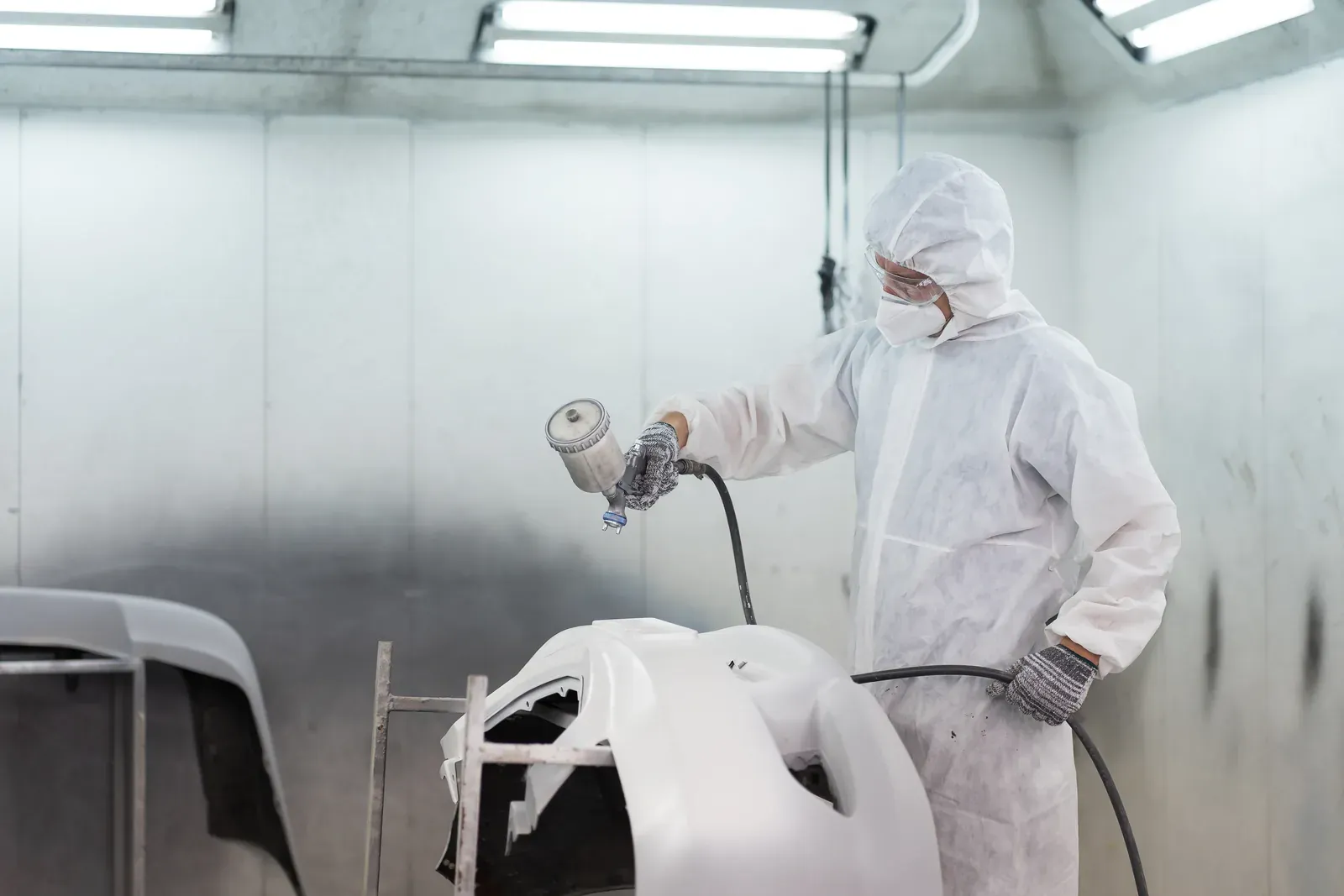 A painter in goggles spraying a white bumper cover on a stand