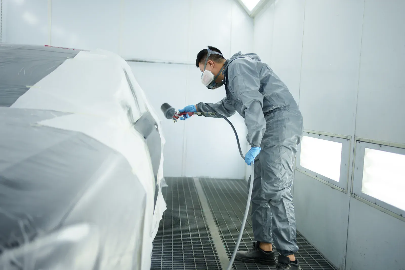 A painter in grey coveralls bending to spray a car masked in white paper